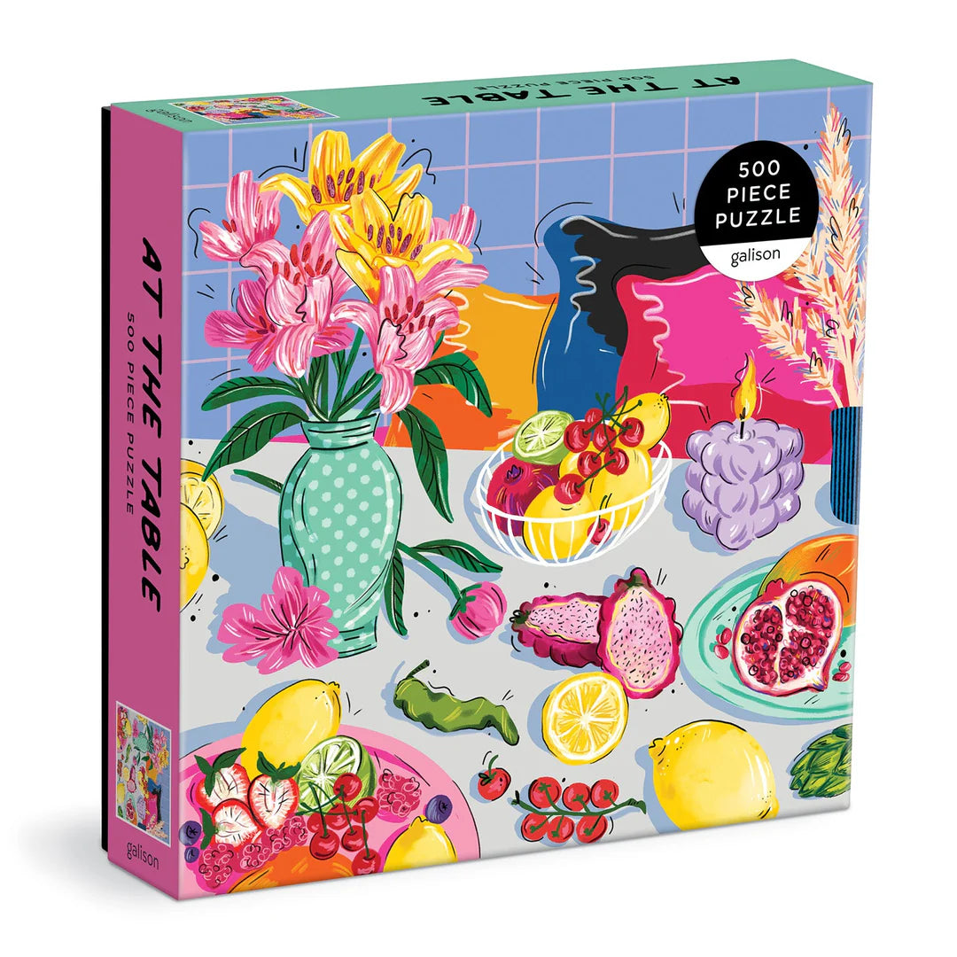 At the Table 500-piece puzzle featuring an illustrated tabletop scene with colorful dishes, cutlery, flowers, and a shared meal setting.